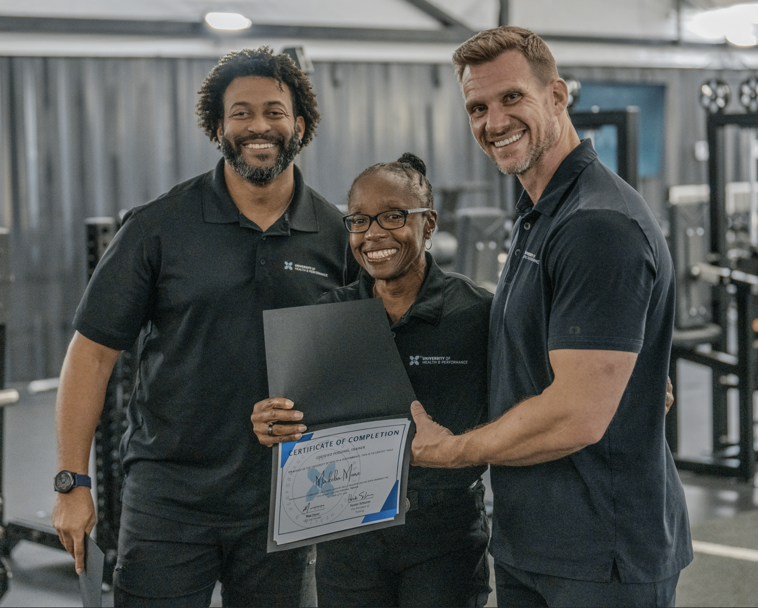 Three people standing together in a gym, smiling. The person in the center holds a certificate of completion, while the other two stand on either side, celebrating the achievement. All are wearing black polo shirts.